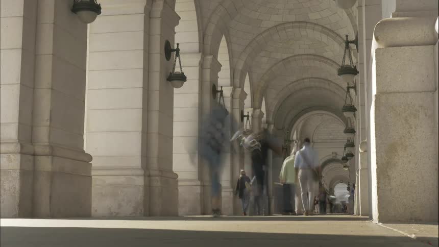 Many people walk the sidewalk of the arched building