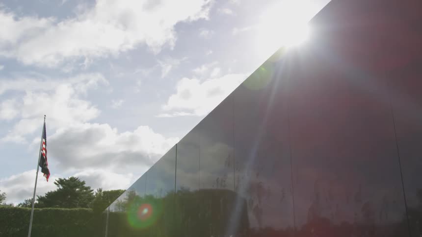 The sun shines over a glossy stone monument as a flag flutters on a pole in the background