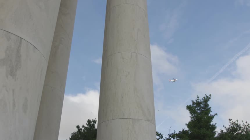 A white column like at lincoln center and the sky is blue with puffy white clouds and airplanes in the distance