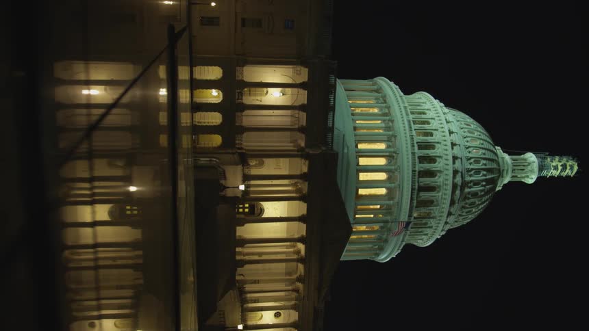 Capitol building during the night with an american flag waving at the front of it