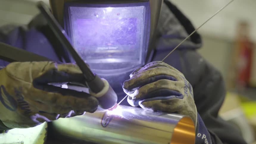 A welder with a hood over the face is welding a piece of metal using gloves and a rod