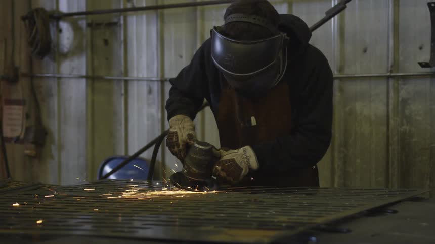 A person in a welding helmet using an electric sander on a sheet of metal