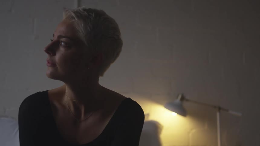 A woman sits on a bed and looks to her right in thought