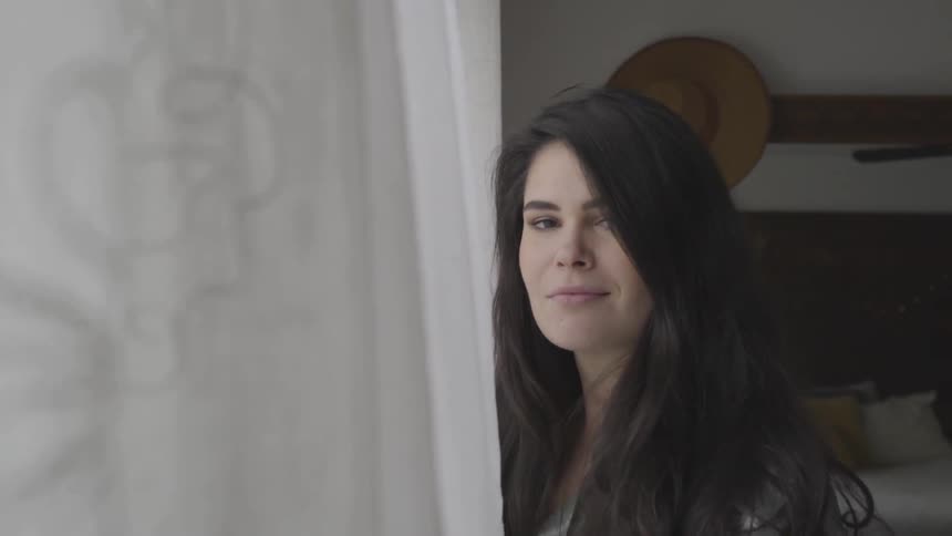 A woman stands by a window and sweeps her hair away from her eyes