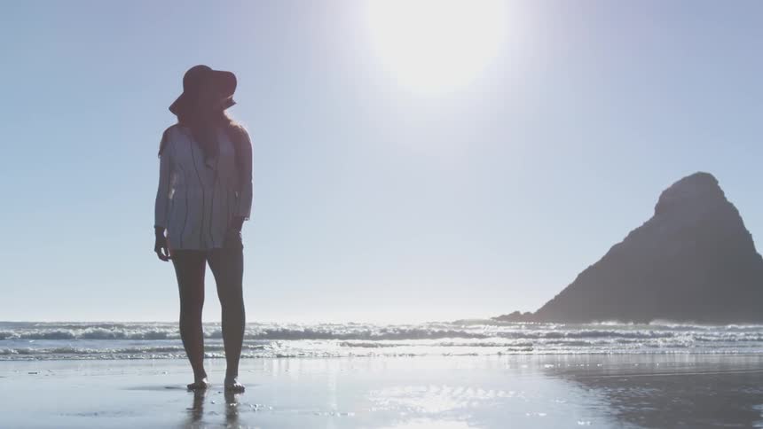 Young woman wearing wide hat stands on golden sand beach next to the sea while the sun shines
