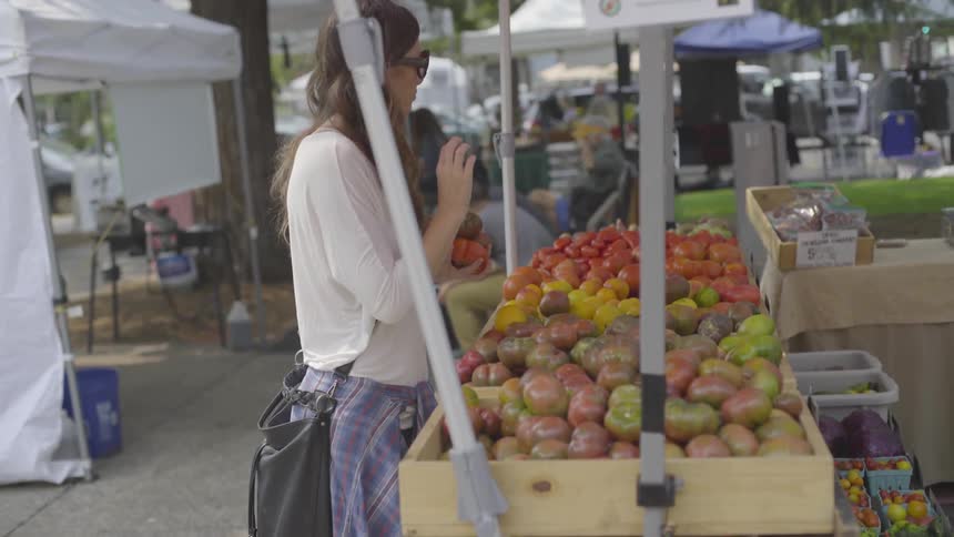 A woman inspecting produce at a farmers market in a city park