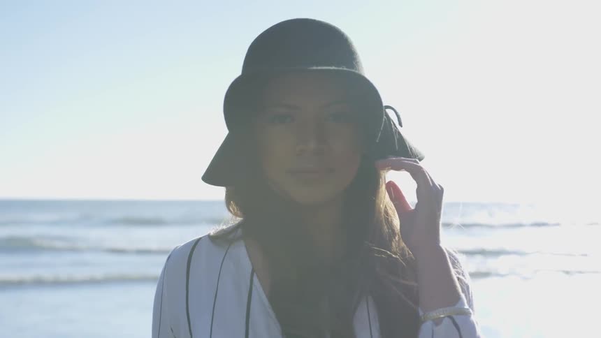 A white young female holding onto her floppy hat standing near a body of water