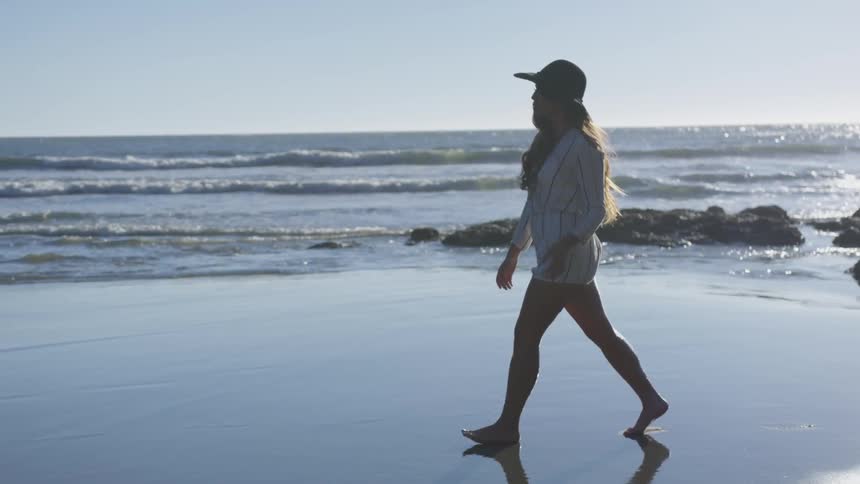 A young woman wearing a sun hat walks barefoot along the beach at sunset