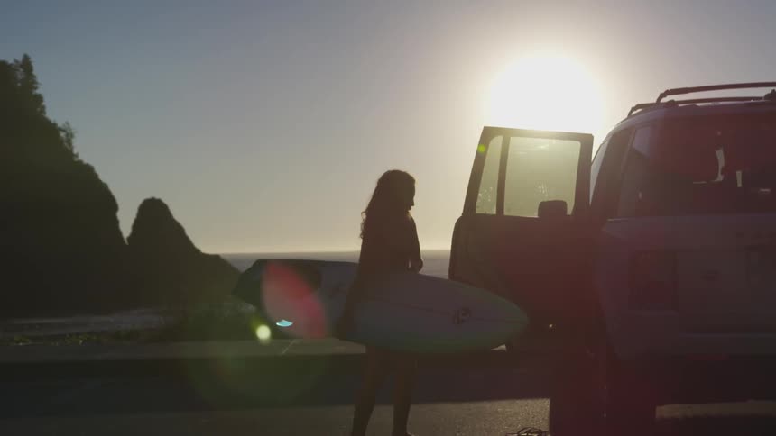 A woman with a surfboard closes a car door at the beach