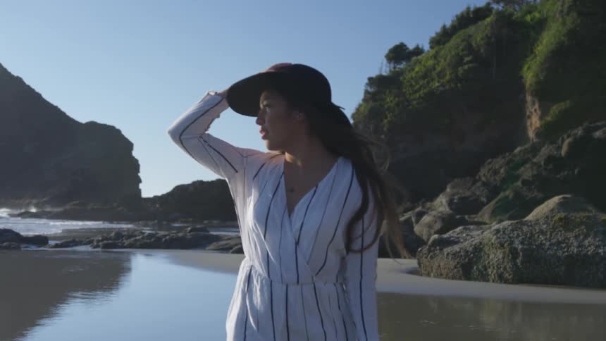 Woman walking at the beach while looking at the ocean and holding her hat