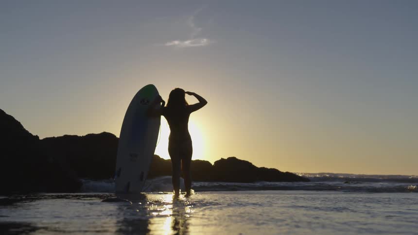 A female surfer is standing in shallow water holding her surfboard while looking out at the waves in the sun