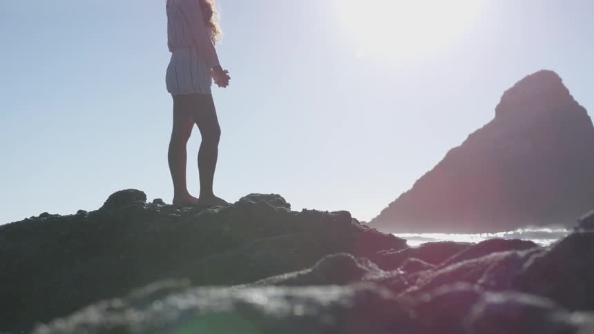 A woman stands on a small rock by the ocean