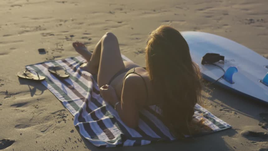 Woman lying on top of a striped towel next to her surfboard at the beach