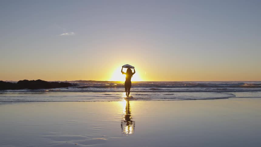 Silhouette of woman with surfboard running towards ocean at sunset