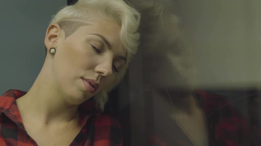 A blonde woman rests her head against the glass pane of an elevator at night