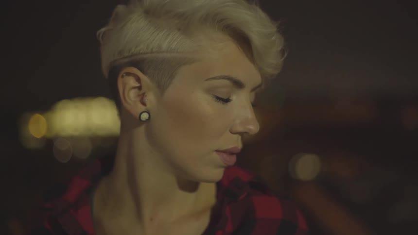 Blonde woman stands with her eyes closed in front of a city horizon at night
