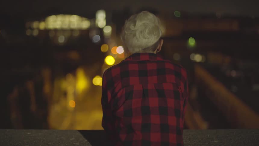 Blond woman who wears striped shirt leans on stone wall with her elbows and look around in city at night
