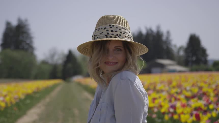 A young woman turns around while walking along a path between two fields of flowers