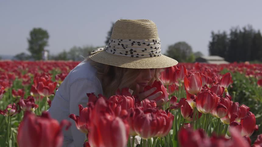A woman squats in a field of red flowers and smells one