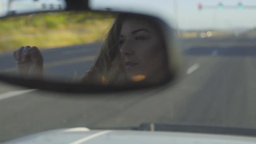 A young woman's face can be seen in the rear view mirror of a car as she brushes her wind blown hair out of her face