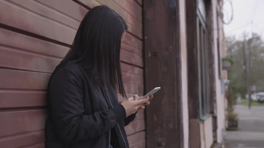 A woman leans against a wall next to a city street and texts on her phone