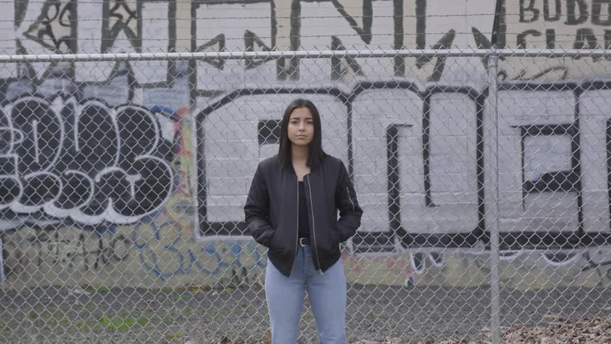 Woman looks into the camera standing in front of a fence and graffiti