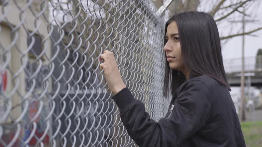 A woman holds on to a chain link fence and looks up at the building behind it