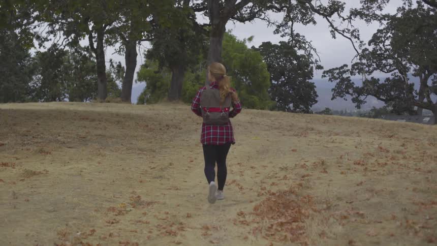 White woman with a backpack walking in the middle of a field towards trees