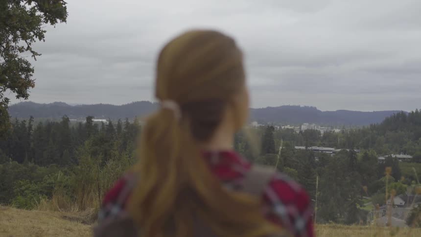 Blonde woman wearing red striped shirt and backpack looks at the countryside and distant mountains in the daytime