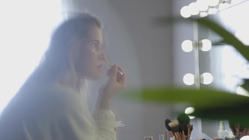 White woman applying makeup in front of an illuminated mirror