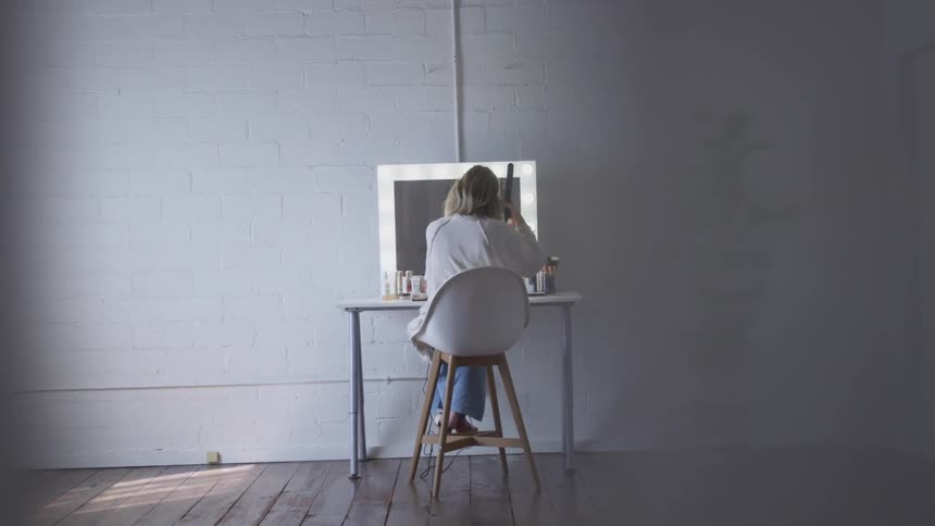 A woman sits at a lit mirrored dressing table using a curling iron