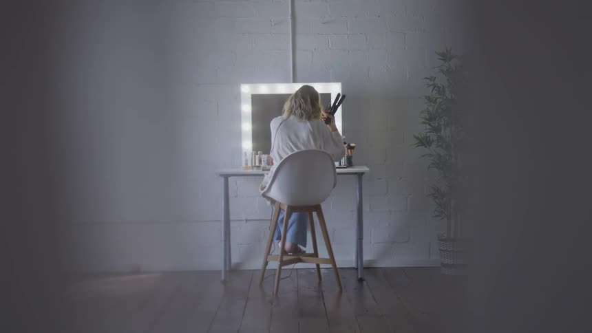 A woman sitting before a vanity curling her hair