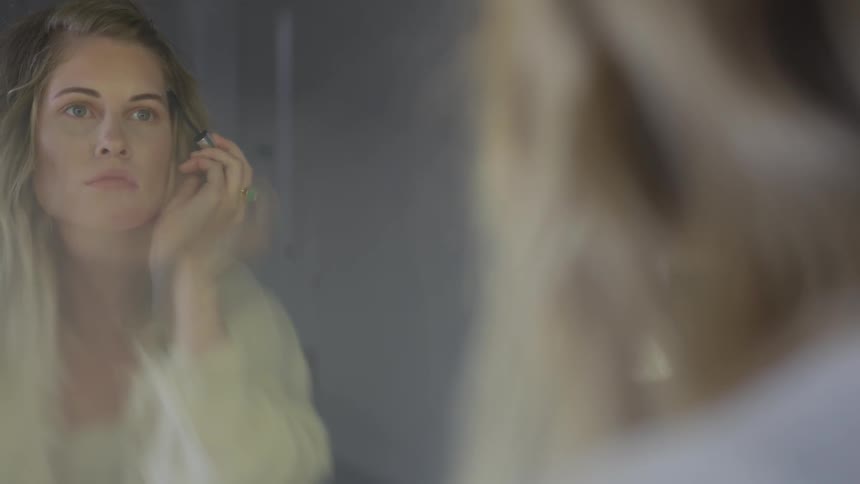 A woman uses a brow spoolie while looking into a vanity mirror