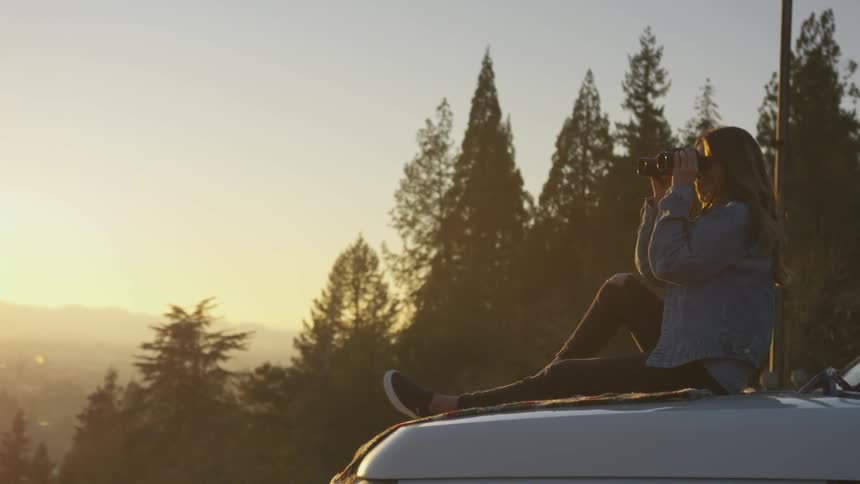 Person uses binoculars to look while sitting on the hood of a car