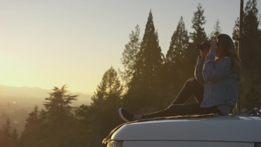 A white woman sits on the hood of a car and looks at the scenery with a pair of binoculars
