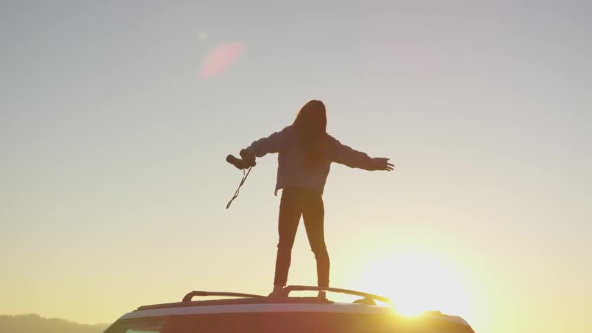 Woman stands on car stretching as sunrises