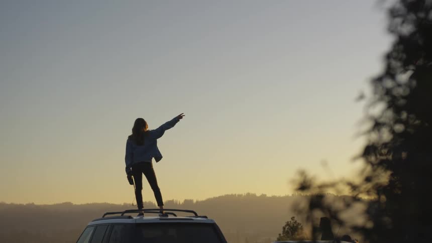 A woman on top of a car pointing a peace sign to the sky