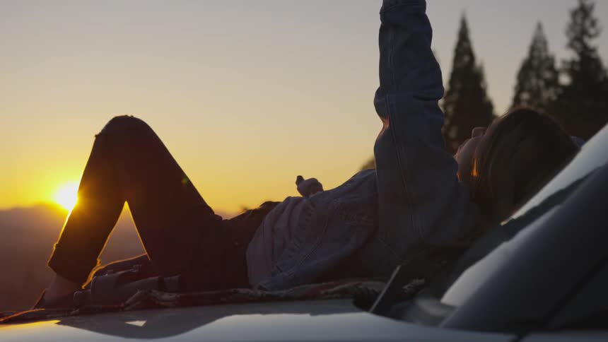 A woman pays on the hood of a car during a sunset