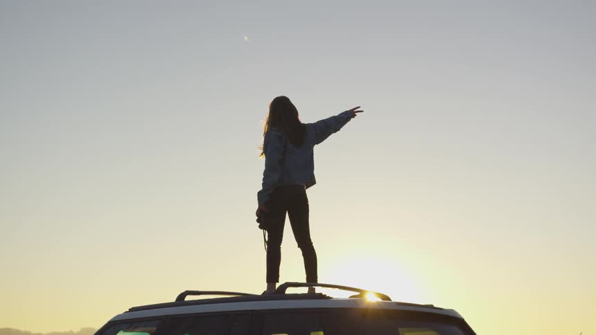 Person stands on top of roof of car at sunset