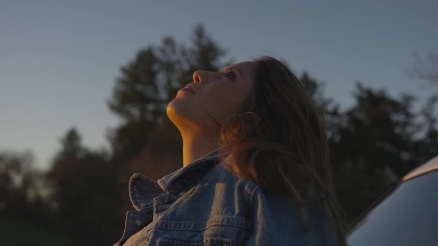 A woman stares into the sky while sitting on the hood of her car