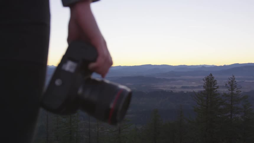 Photographer stands looking down at forest scenery while holding a camera