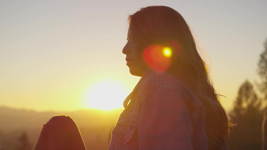 Woman with long hair and denim jacket sits down and look ahead in the forest at sunset