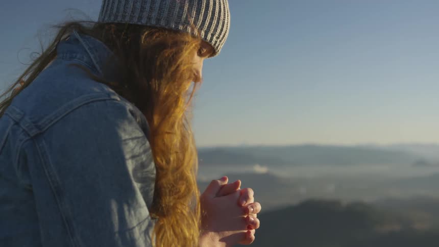 White woman praying while standing at the top of a mountain