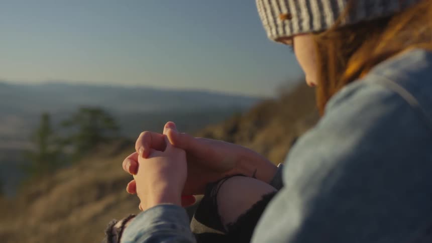 Woman sits with bowed head and clasped hands on slope over valley