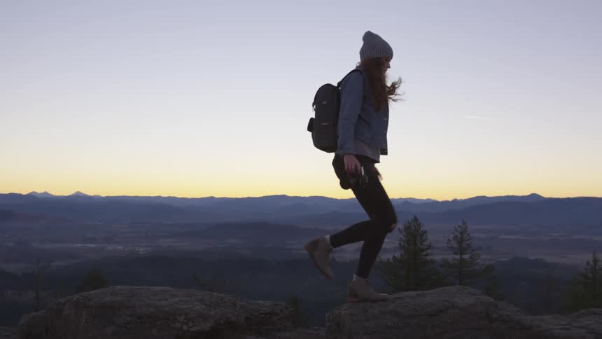 Woman carrying professional camera jumps from one rock to another at dusk