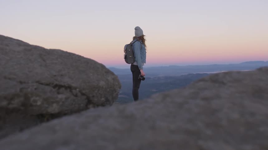 A woman with a backpack and camera stands on a cliff looking at the horizon