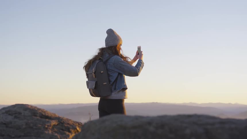 Woman with a backpack filming something with her phone while standing on a hill