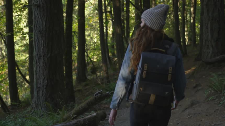 Woman wearing wooly hat and backpack takes picture of trees with smart phone in the forest in the daytime