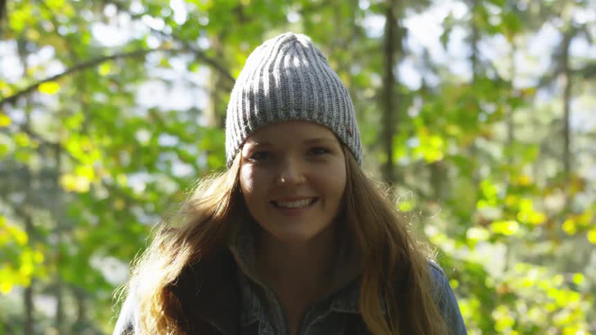 A young woman wearing a beanie smiles while in a wooded area