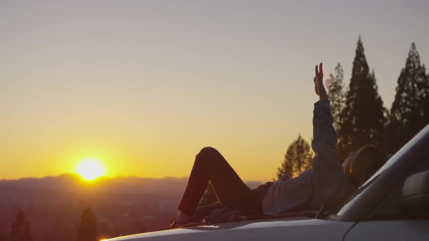 A man lays on his hood raising his hand to a beautiful sunrise